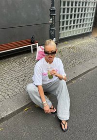 Λευκό oversized t-shirt με ροζ κείμενο, γκρι φαρδύ παντελόνι φόρμας, μαύρα σάνταλα, ασημένια βραχιόλια και ένα πράσινο ροφημα σε διάφανο ποτήρι.