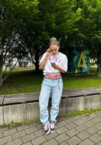 Weißes T-Shirt mit pinkem "NIKE" Logo, hellblaue, weite Jeans, grau-schwarze Sneakers mit pinken Schnürsenkeln. Person, die draußen ein Smartphone benutzt.