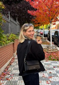 Must ristperkott klapi ja hõbedase riistvaraga, kantud musta jakiga. Naine hoiab kohvitassi ja kõnnib lehtedega kaetud rajal.