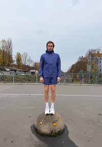 Giacca sportiva viola e pantaloncini, in piedi su una pietra arrotondata con accenti arancioni. Sullo sfondo edifici e alberi sotto un cielo nuvoloso.