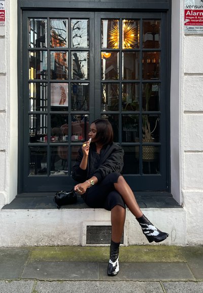 Chaqueta bomber negra, falda negra ajustada y botas de tobillo en texturas en blanco y negro. Sentada en un borde de piedra, sosteniendo comida. Puerta de cristal en el fondo.