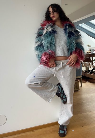 Chaqueta de pelo sintético colorida con hebras rosas, azules y blancas, top gris corto, pantalones deportivos grises y zapatos deportivos multicolores.