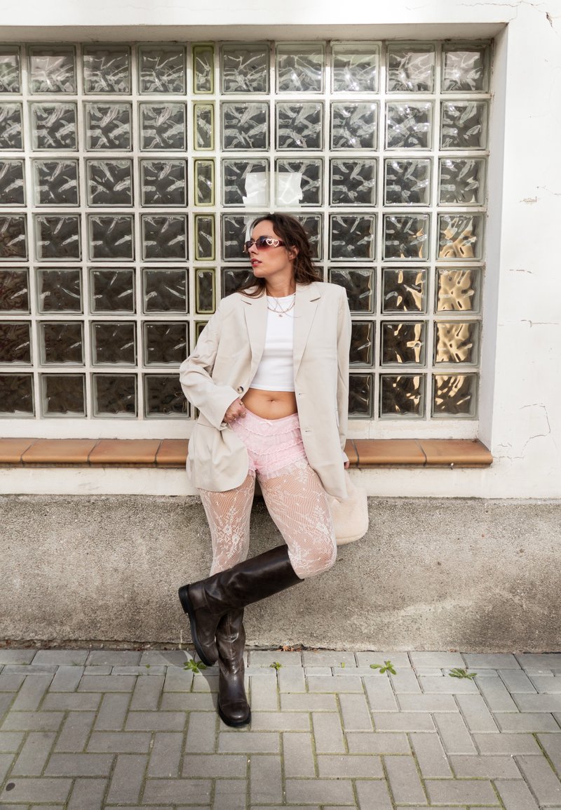 Blazer oversized bege, top cropped branco, calções de renda rosa, meias-calças estampadas e botas pretas até ao joelho, contra uma parede de vidro texturizado.