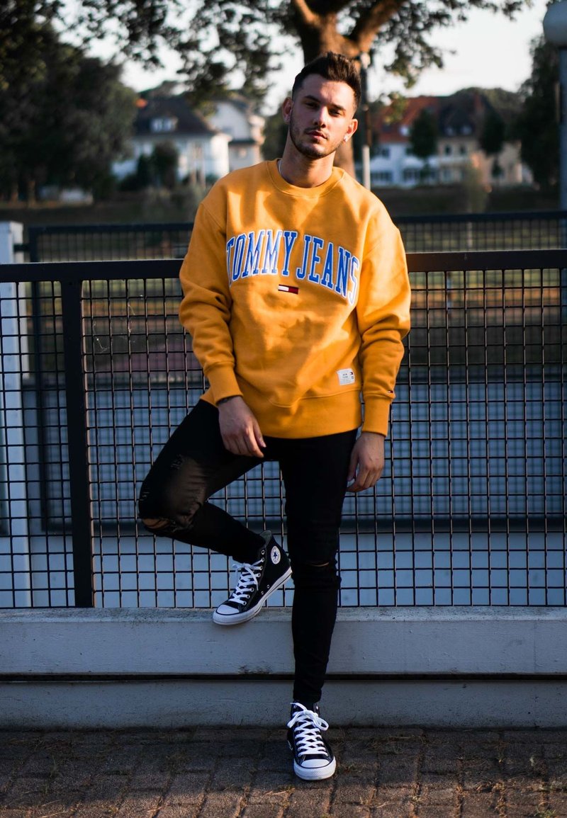 Gelbes Sweatshirt mit "TOMMY JEANS" in blauer Schrift, schwarze zerrissene Jeans und schwarze High-Top-Sneaker mit weißen Akzenten, die an einem Zaun stehen.