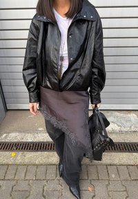 Czarna skórzana kurtka typu bomber, szara przezroczysta warstwowa spódnica z koronkowym wykończeniem, czarne dżinsy, spiczaste buty i teksturowana czarna torebka.