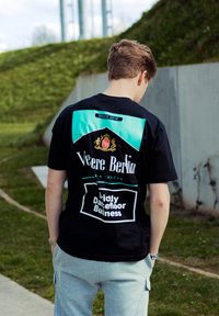 Juodas marškinėliai su turkio akcentais ir balta tekstu. Yra „Veere Berlin“ logotipas, „Nuo 2016“ ir dizainas „Griežtai šokių aikštelės verslas“. 
