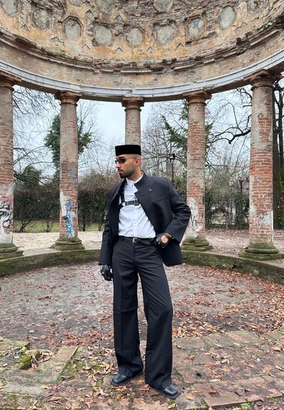 Hombre con traje negro, camisa blanca, gafas de sol y sombrero posa bajo una ruina de piedra circular con altas columnas de ladrillo al aire libre.