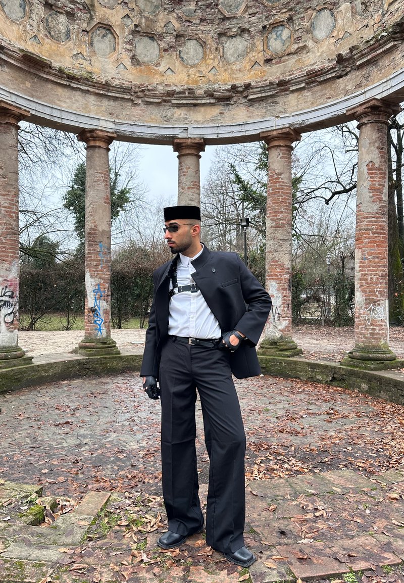 Homme en costume noir, chemise blanche, lunettes de soleil et chapeau pose sous une ruine circulaire en pierre avec de hautes colonnes en brique en plein air.