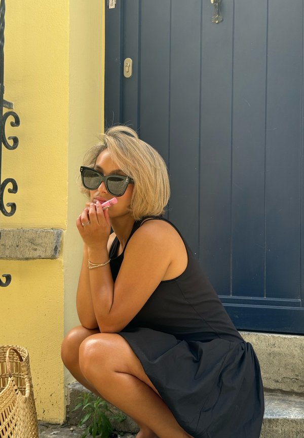Abito nero senza maniche, occhiali da sole neri oversize, capelli castano chiaro, tiene un prodotto per le labbra rosa, accovacciata contro un muro giallo e una porta blu.