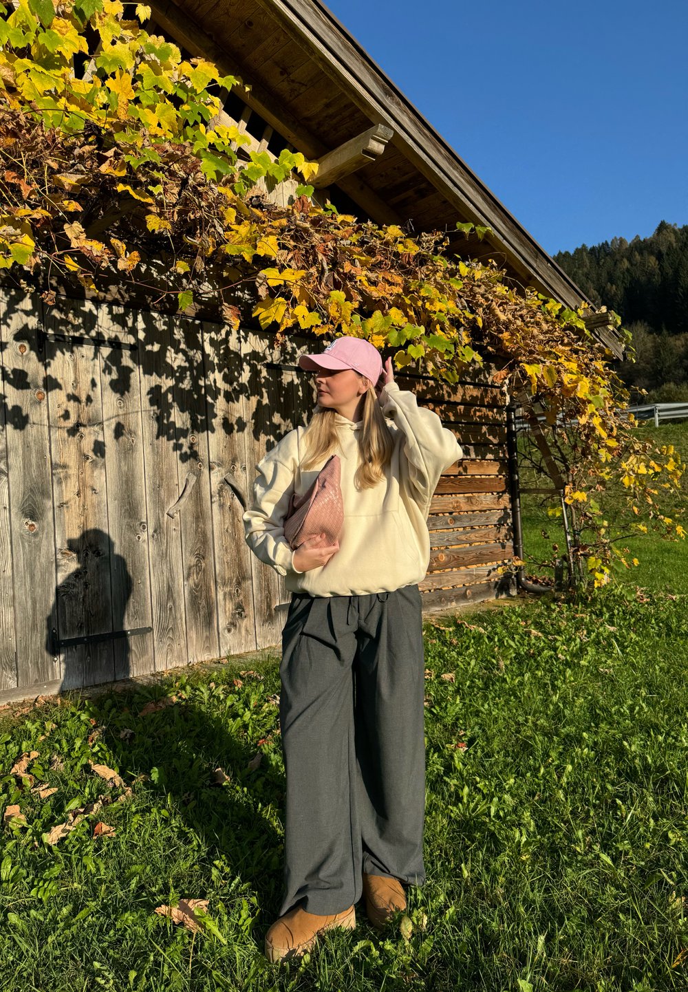 Sweatshirt oversized em creme, calças largas cinzentas e boné rosa. Segura uma clutch texturizada rosa. O fundo apresenta uma parede de madeira e folhas de outono.