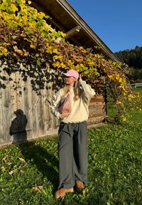 Sweatshirt oversized em creme, calças largas cinzentas e boné rosa. Segura uma clutch texturizada rosa. O fundo apresenta uma parede de madeira e folhas de outono.
