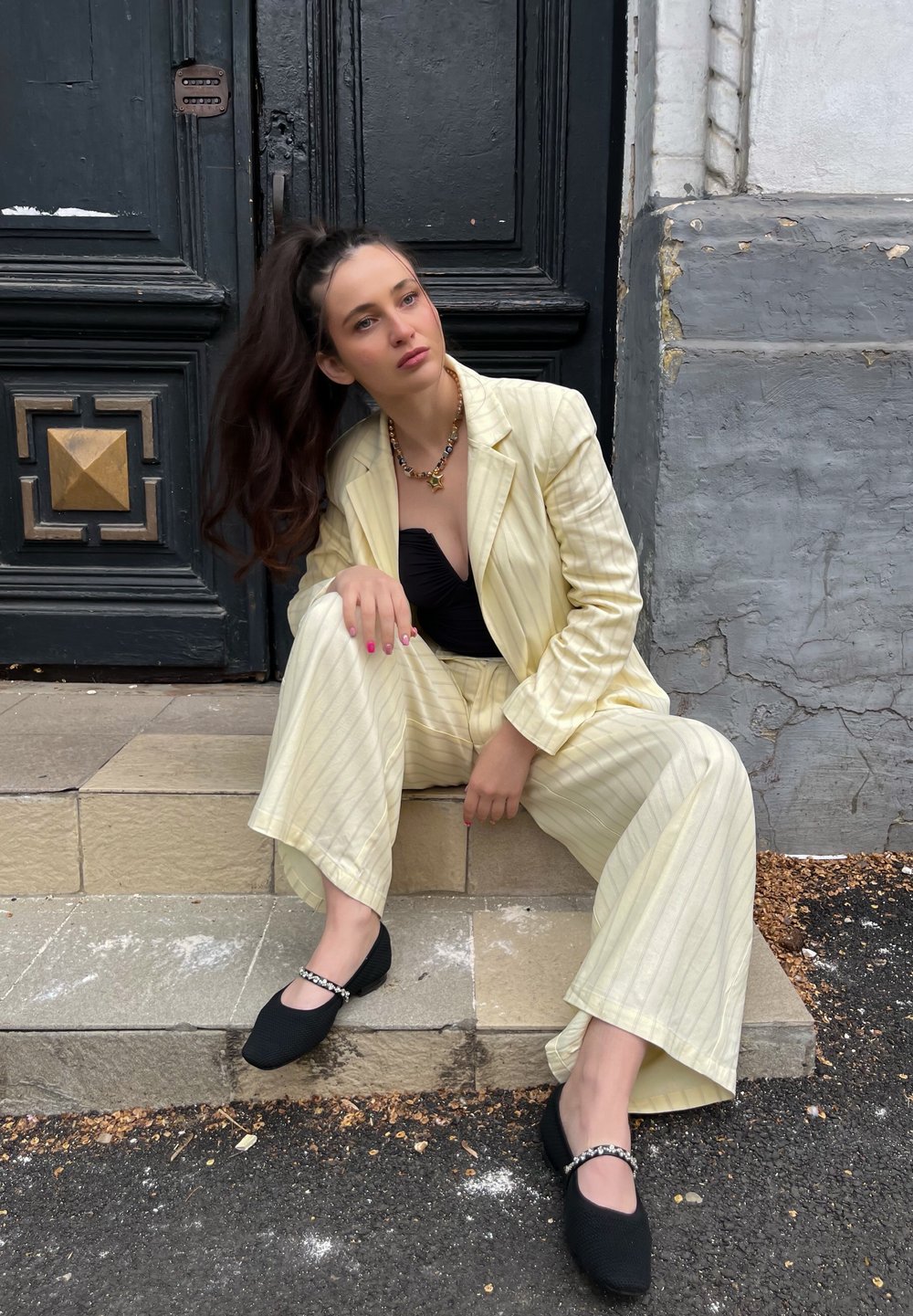 Ανοιχτό κίτρινο κοστούμι με ρίγες και φαρδιές παντελόνες, σε συνδυασμό με μαύρο τοπ και μαύρες υ�φασμάτινες μπαλαρίνες με αλυσίδα ως διακόσμηση.