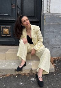 Ανοιχτό κίτρινο κοστούμι με ρίγες και φαρδιές παντελόνες, σε συνδυασμό με μαύρο τοπ και μαύρες υφασμάτινες μπαλαρίνες με αλυσίδα ως διακόσμηση.