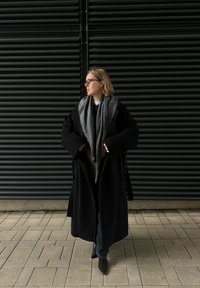 Black oversized coat with wide sleeves, layered over a grey scarf. Model stands on a tiled surface, with a dark louvered wall in the background.