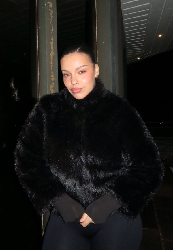 Kobieta z gładko zaczesanymi włosami w czarnej, puszystej kurtce, stoi nocą z rękami złożonymi, przy metalowych belkach i oświetleniu sufitowym.