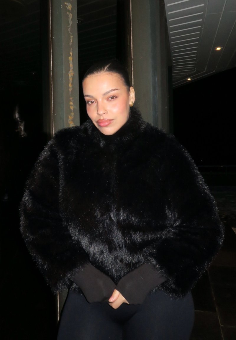 Kvinne med glatt bakoverstrøket hår iført svart pelsjakke, står med hendene foldet om natten nær metallbjelker og taklys.