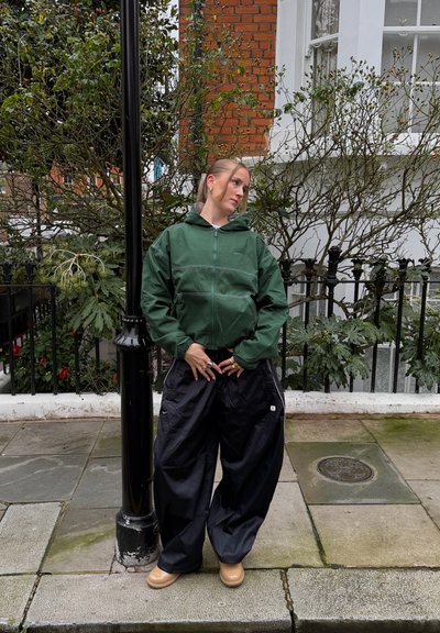 Persona con chaqueta verde con cremallera y pantalones anchos negros apoyada en un farol negro en la acera, con plantas y un edificio de ladrillo detrás.