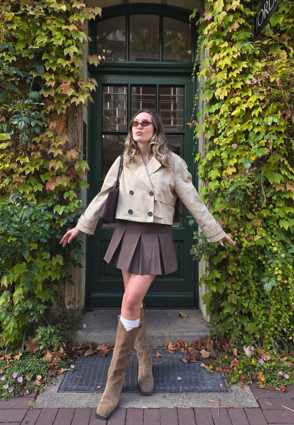 Beige cropped jacket, brown pleated skirt, tan cowboy boots with a textured design. The background features green ivy and a dark green door.