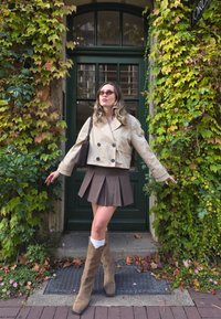Beige cropped jacket, brown pleated skirt, tan cowboy boots with a textured design. The background features green ivy and a dark green door.
