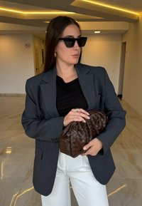 Sivý károvaný blejzer, čierne tričko, biele nohavice, veľké čierne slnečné okuliare, držiac hnedé pletené psaníčko. Hladké textúry, štruktúrovaný dizajn.