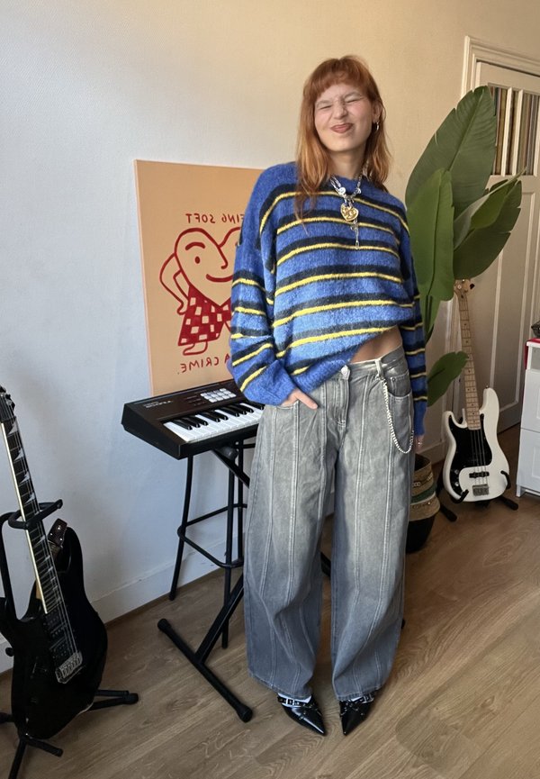 Osoba s červenými vlasmi, oblečená v modro-žltom pruhovanom svetri a širokých sivých nohaviciach, stojí usmievajúca sa pri keyboarde, gitarách a veľkej zelenej rastline.