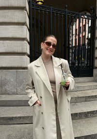 Femme en manteau beige et lunettes de soleil tenant une boisson verte avec une paille, debout près d'un escalier en pierre et d'une grille en fer forgé noire.