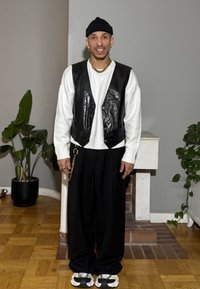 Gilet in pelle nera sopra un maglione bianco, pantaloni neri e sneaker con fantasia. Accessori includono una catena d'oro e orecchini. Ambientazione interna.