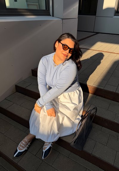 Mujer con gafas de sol, camiseta azul de manga larga, falda blanca y bailarinas en blanco y negro, sentada en unas escaleras exteriores con una bolsa negra a su lado.