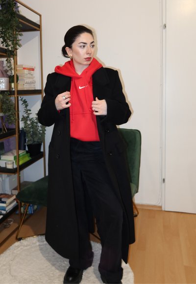 Abrigo de lana negro sobre una sudadera con capucha roja de Nike, con pantalones oscuros y botas negras. El atuendo incluye anillos y una estantería de pared con textura en el fondo.