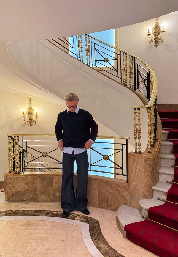 Mężczyzna poprawia sweter stojąc przy krętych schodach z czerwonym dywanem i ozdobną złotą balustradą w eleganckim wnętrzu.