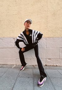 Czarny i biały paskowany dres Adidas, czarne spodnie z białymi paskami i fioletowe buty Nike. Model pozuje przy teksturowanej ścianie.