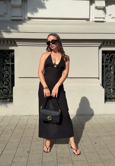 Vestido maxi halter negro con un lazo en el frente, tirantes finos y una textura transparente, combinado con un bolso negro y chanclas sobre una superficie pavimentada.