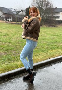 Jeune femme en jeans bleu clair, veste en peau retournée marron et bottes foncées, souriant debout sur un trottoir mouillé près d'une zone herbeuse.