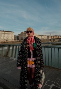 Cappotto oversize nero e rosa con motivo a fantasia, maglione rosa acceso con dettagli floreali, gonna rosa e borsa bianca. Occhiali da sole e sciarpa completano il look.