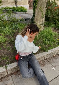 Maglione cropped bianco indossato sopra una maglietta a fantasia floreale, abbinato a jeans grigi scuri. Gli accessori includono orecchini rossi e una piccola borsa rossa.