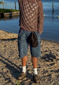 Pull en tricot marron avec un motif en diamant, short en denim bleu, chaussettes blanches, chaussures brunes et une casquette noire à la main, se tenant sur le sable près de l'eau.