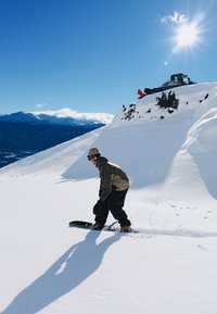 Snowboardeur en tenue marron et noire sur une pente enneigée. Ciel bleu vif avec soleil et montagnes lointaines en arrière-plan. Chasse-neige sur la crête.