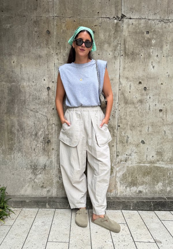 Ung kvinde iført grønt tørklæde, sorte solbriller, grå ærmeløs top, beige vidde bukser og beige slip-on sko, står op ad en betonvæg.
