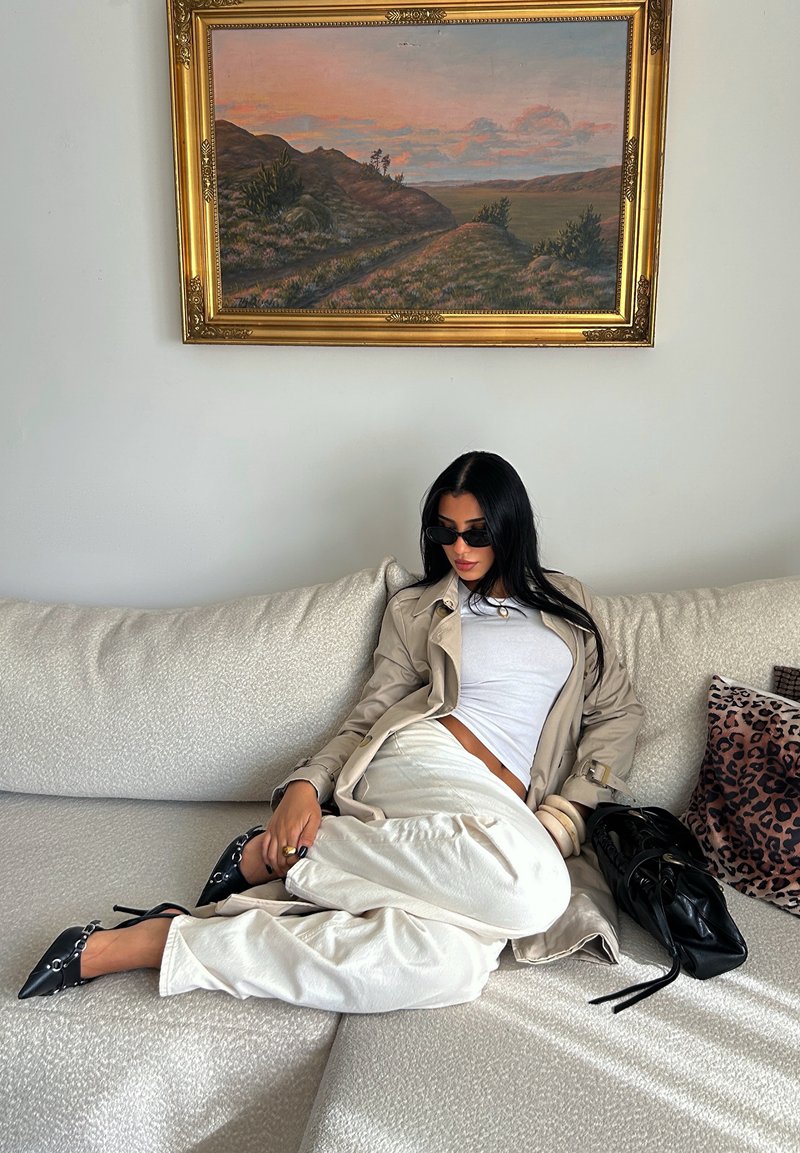 Cappotto beige sopra una maglietta bianca e pantaloni crema, seduta su un divano chiaro con texture. Tacchi neri e una borsa nera, con un cuscino con motivo.