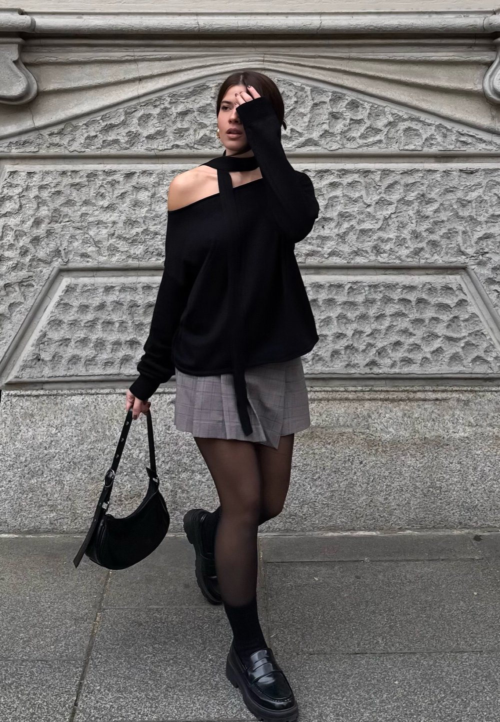 Μαύρος off-shoulder πουλόβερ, γκρι καρό μίνι φούστα, μαύρες καλσόν, χοντροί μαύροι loafers και μια μικρή μαύρη τσάντα, τοποθετημένα μπρο στά από έναν υφασμάτινο τοίχο.