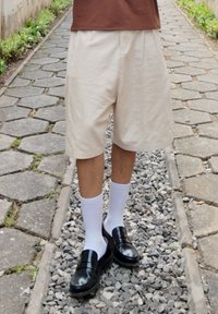 Short beige avec plis, porté avec des chaussettes blanches montantes et des mocassins noirs brillants. En arrière-plan, un chemin en pierre bordé de verdure.