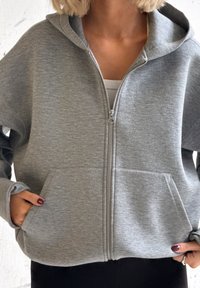 Osoba ubrana w jasnoszarą bluzę z kapturem i suwakiem, trzymająca ręce w przednich kieszeniach, pod spodem biała koszula, a u dołu widoczne czarne spodnie.