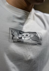 Hvit bomulls t-skjorte med et svart-hvitt grafisk trykk som viser en karakter med store ører og uttrykksfulle øyne, satt mot en teksturert bakgrunn.