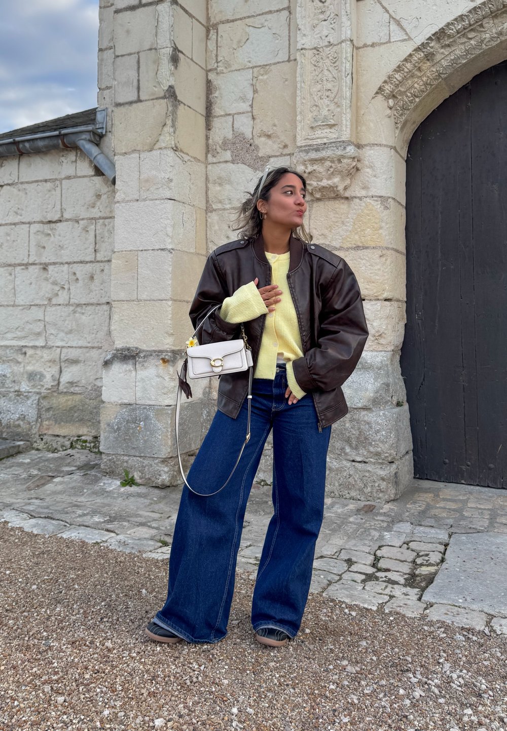 Brown leather bomber jacket over a light yellow jumper, teamed with wide-leg blue jeans and a small white handbag with a chain strap.