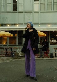 Manteau en fausse fourrure noire sur un costume violet ; pantalon large et élégant ; cheveux courts avec des reflets bleus et violets ; café urbain en toile de fond avec des sièges en plein air.