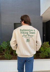 Kremowa bluza z zielonym napisem "Drużyna Badmintona i Wędrówek - Dom Klubu" oraz różanym wzorem na plecach, w połączeniu z niebieskimi dżinsami.