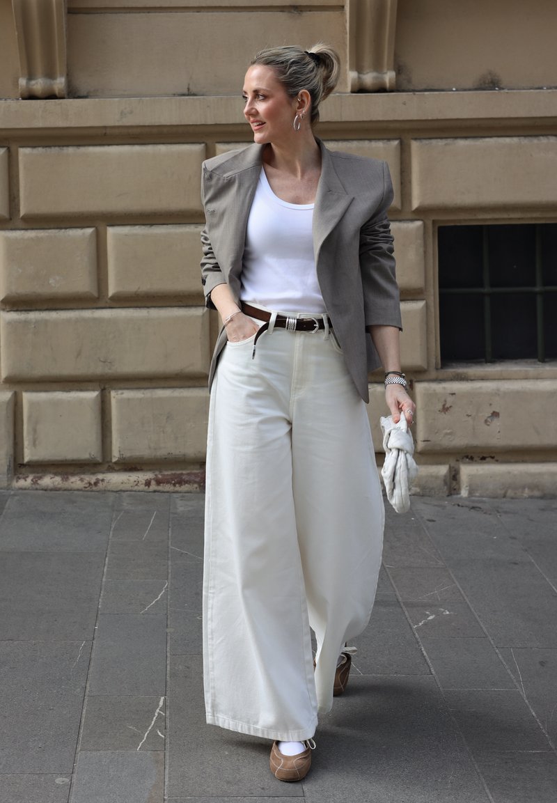 Woman walking on city sidewalk wearing a gray blazer, white tank top, wide white pants, and brown shoes, holding a scrunched cloth in hand.