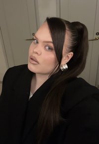 Abrigo negro con cuello alto, llevado sobre una blusa blanca. Cabello largo y castaño peinado en una coleta. Gran aro de plata en la oreja izquierda.