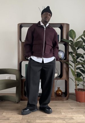 Chaqueta granate con cremallera y dos bolsillos delanteros, camisa blanca debajo, pantalones negros y zapatos negros gruesos. Ambiente interior con plantas.