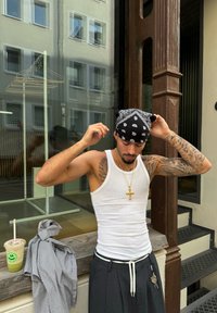 Homme ajustant un bandana noir, portant un débardeur blanc et un pantalon large gris avec un cordon blanc, affichant des tatouages et une chaîne en or avec une croix.